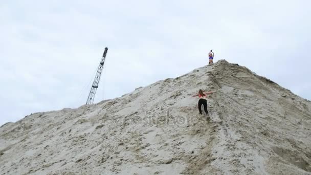 Junge Sportlerin in rotem Top und schwarzer Leggings erklimmt einen sandigen Berg. Im Hintergrund sieht man Frachtkräne, Frachthafen, Morgendämmerung. — Stockvideo