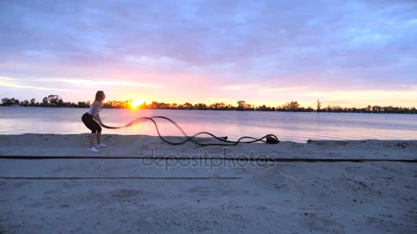 Mujer joven, atlética, rubia, realiza ejercicios de fuerza con la ayuda de una cuerda gruesa y deportiva. Al amanecer, en la playa de la cantera de arena, a la luz del sol, video rodaje contra el sol . — Vídeos de Stock