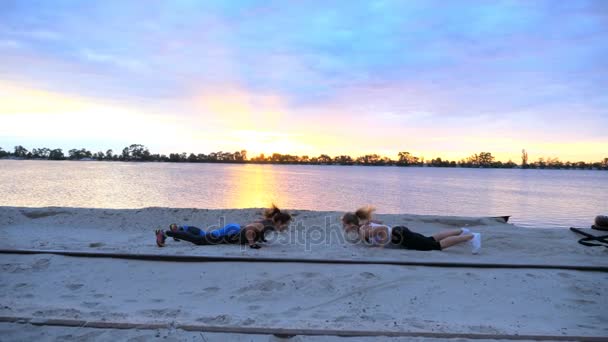 Mooie, slanke jonge vrouwen zijn bezig met fitness, springen, kracht oefeningen, push-ups uit te voeren. Bij zonsopgang, zonsopgang, op het zandstrand steengroeve strand, video schieten tegen de zon. — Stockvideo