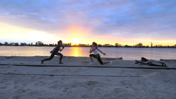 Schöne, schlanke junge Frauen treiben Fitness, springen, machen Kraftübungen, Liegestütze. im Morgengrauen, Sonnenaufgang, am Sandstrand des Steinbruchs, Videodreh gegen die Sonne. — Stockvideo