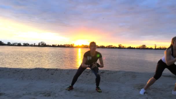 漂亮、 苗条的年轻妇女从事健身、 跳转、 执行力量练习，俯卧撑。在黎明，日出，海边沙地采石场、 视频拍摄对着太阳. — 图库视频影像