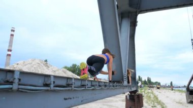 Mavi bir üst ve çizgili leggens atletik kadın gücü çalışmaları gerçekleştirir. O bir yük vinç metal kiriş üzerinde duruyor. bir kargo liman plajında şafak, Fitness.