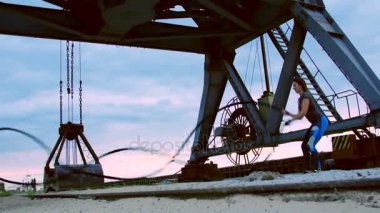 Genç, atletik, kadın kalın ve sportif bir iple güç egzersizleri yapıyor. Şafakta, kargo limanının kumsalında, güneş ışığında, bir kargo vincinin yanında.,