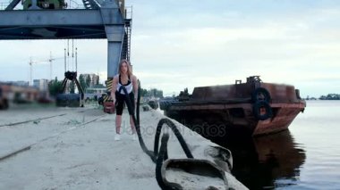 Genç, atletik, kadın kalın ve sportif bir iple güç egzersizleri yapıyor. Şafakta, kargo limanının kumsalında, güneş ışığında, bir kargo vincinin yanında.,