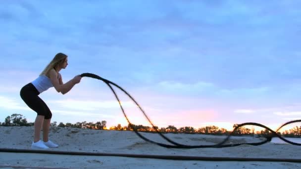 Joven, atlética, mujer, realiza ejercicios de fuerza con la ayuda de una cuerda gruesa y deportiva. Al amanecer, a lo largo del muelle de arena del puerto de carga, a la luz del sol, video rodaje contra el sol . — Vídeos de Stock