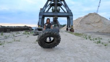 Gün doğumunda, Genç bir atletik kadın büyük bir ağır traktör tekerleği kullanarak egzersizleri yapar, onu kasları antrenörler. Atıyor. Arka planda kargo vinçler, kargo limanı, şafak görülebilir.