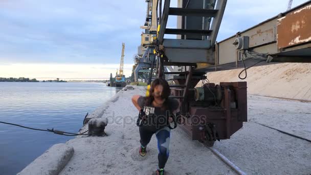 Beautiful lady in T-shirt, blue leggens exercising her muscles with help of suspension straps TRX. At dawn, sunrise, near a cargo crane, on the sandy quarry beach, near the river, — Stock Video