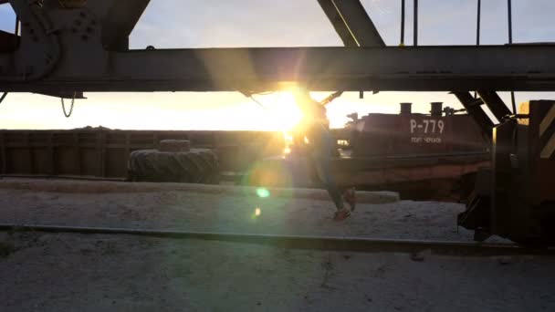 Красивая леди тренирует мышцы с помощью подвесных ремней TRX. На рассвете, в лучах солнца, рядом с грузовым краном, на песчаном карьерном пляже, рядом с рекой , — стоковое видео