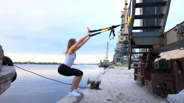Красива леді тренує м'язи за допомогою суспензійних ременів TRX. На задньому плані видно річку, вантажні крани, вантажний порт. На світанку, на піщаному кар'єрному пляжі . — стокове відео