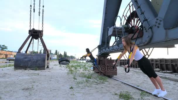 Bella donna atletica che fa esercizio con sistema trx. Lei fa flessioni braccia di formazione con cinghie fitness trx. All'alba, vicino a una gru da carico, sulla spiaggia sabbiosa della cava , — Video Stock