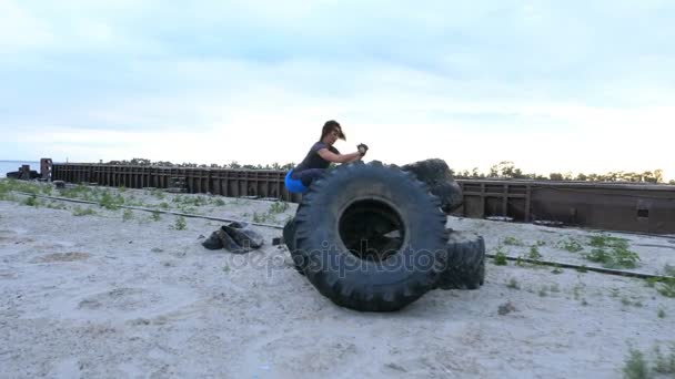 Атлетическая женщина в футболке, синие леггенсы выполняет силовые упражнения с помощью большого колеса трактора, она прыгает. На пляже, в грузовом порту, на песке. Рассвет, на рассвете. Медленное движение. — стоковое видео