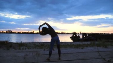 Güzel, ince genç kadın gerçekleştirir güç egzersizleri fitness konusunda meşgul. Şafakta güneşin karşı çekim video kargo bağlantı noktası, kum iskele boyunca. Güneş doğarken,