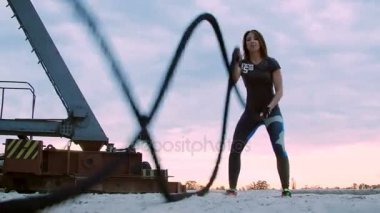 Genç, atletik, kadın kalın ve sportif bir iple güç egzersizleri yapıyor. Şafakta, kargo limanının kumsalında, güneş ışığında, bir kargo vincinin yanında.,