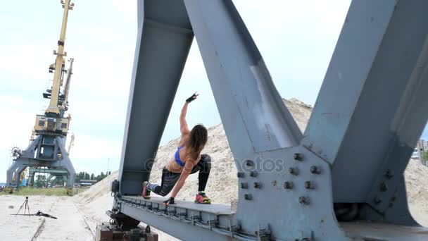 Mujer atlética en una parte superior azul y las piernas rayadas realiza ejercicios de fuerza.Ella está parada en el haz de metal de una grúa de carga. fitness al amanecer, en la playa en un puerto de carga . — Vídeos de Stock