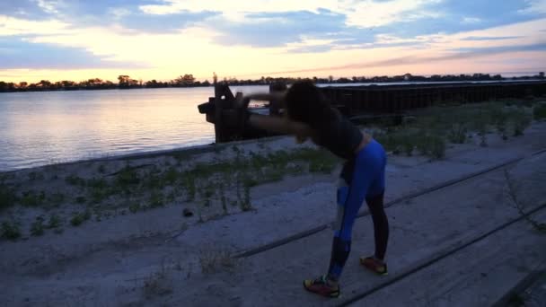 Mooie, slanke jonge vrouw houdt zich bezig met fitness, voert kracht oefeningen. Bij dageraad, langs de zand pier van de vrachthaven, video schieten tegen de zon. Bij zonsopgang, — Stockvideo