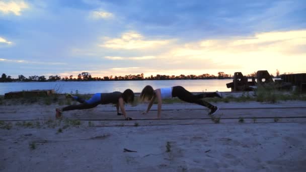 Mooie, slanke jonge vrouwen zijn bezig met fitness, kracht oefeningen uit te voeren. Bij dageraad, langs de zand pier van de vrachthaven, video schieten tegen de zon. Bij zonsopgang, — Stockvideo