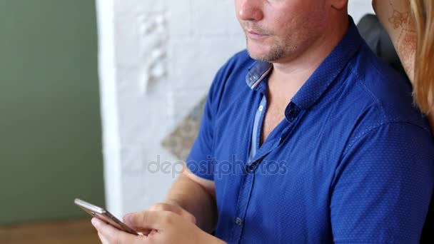 Um homem caucasiano está segurando um telefone, um smartphone, olhando para ele, digitando texto, enquanto menina, mestre de tatuagem, artista mehendi faz desenho de tatuagem de hena em seu couro cabeludo, cabeça — Vídeo de Stock