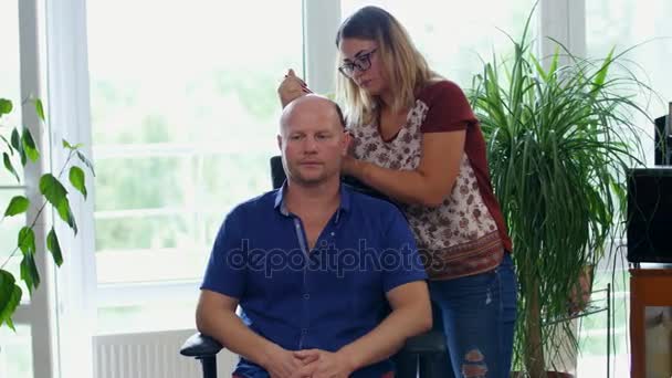 Uma menina, um mestre, um artista mehendi, uma tatuagem, faz um desenho de hena no couro cabeludo, um homem caucasiano careca, em uma camisa azul, sentado em uma cadeira.Mehendi, tatuagens de hena, agora é muito popular entre os homens. — Vídeo de Stock