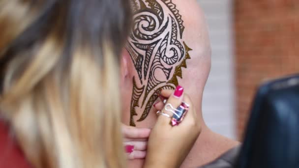 En flicka, tatuering master, mehendi artist gör ritning av henna tatuering på hårbotten skallig kaukasiska man, axel, nacke. Processen att tillämpa henna från ett rör på huden. — Stockvideo