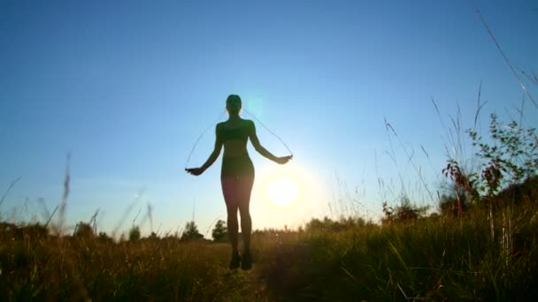 Figura, contorno de una hermosa chica atlética con el pelo largo y rubio, saltando la cuerda al atardecer, contra el cielo azul, a la luz del sol, en verano . — Vídeo de stock