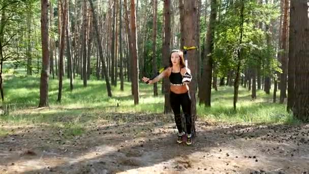 Mulher bonita, atlética, sexy jovem, treinador, instrutor, realiza exercícios, fazendo exercícios com sistema de fitness trx, correias de suspensão TRX. Na floresta de pinheiros, no verão, nos raios de sol . — Vídeo de Stock