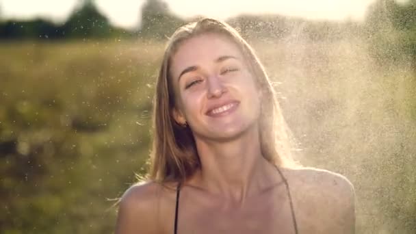 Portrait of a beautiful young blonde girl, under light summer rain, in the sun rays, on a green meadow. The girl smiles, strokes herself over the delicate, beautiful white skin. — Stock Video