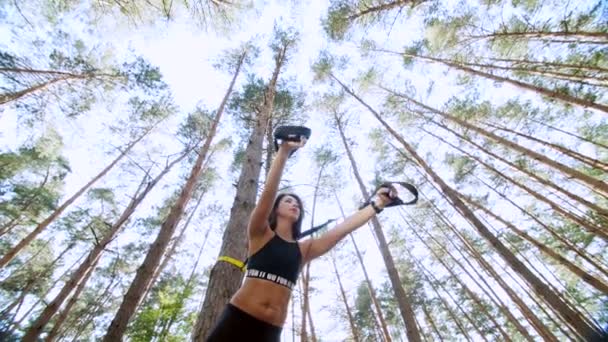 Mujer atlética, entrenador, instructor, realiza, haciendo ejercicios con sistema de trx fitness, correas de suspensión TRX. En el bosque de pinos, en verano, en los rayos del sol. Vista desde abajo, vista inferior — Vídeo de stock