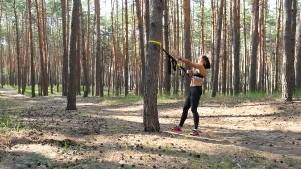 Beautiful, athletic, sexy young woman, coach, instructor, performs exercises, doing exercises with fitness trx system, TRX suspension straps. In pine forest, in summer, in sun rays. — Stock Video