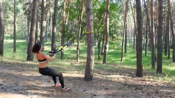 Belle, athlétique, sexy jeune femme, entraîneur, instructeur, effectue des exercices, faire des exercices avec le système de fitness trx, sangles de suspension TRX. En forêt de pins, en été, sous les rayons du soleil .