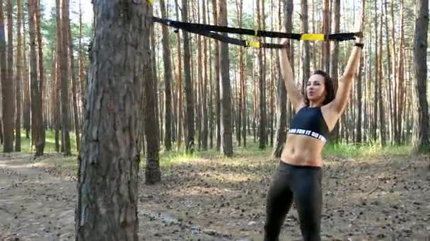 Mulher bonita, atlética, sexy jovem, treinador, instrutor, realiza exercícios, fazendo exercícios com sistema de fitness trx, correias de suspensão TRX. Na floresta de pinheiros, no verão, nos raios de sol . — Vídeo de Stock