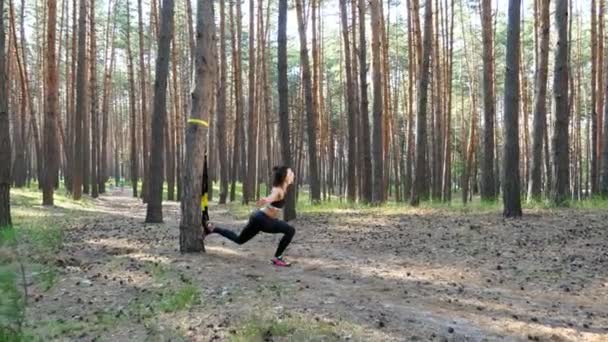 Hermosa, atlética, mujer joven sexy, entrenador, instructor, realiza ejercicios, haciendo ejercicios con sistema de trx fitness, correas de suspensión TRX. En el bosque de pinos, en verano, en los rayos del sol . — Vídeo de stock