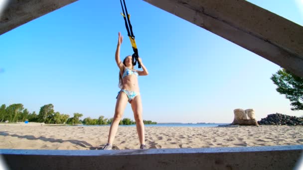 Belle, athlétique, sexy jeune femme en maillot de bain, entraîneur, instructeur, effectue, faire des exercices avec le système de fitness trx, sangles de suspension TRX. Sur la plage, en été, sous les rayons du soleil . — Video