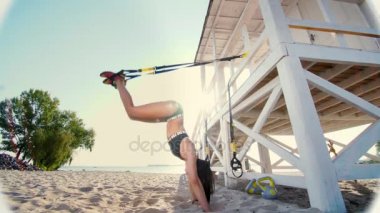 atletik, seksi mayo, genç bir kadın duruyor ellerinde, baş aşağı, fitness trx sistemi, Trx askı kayışları ile yapıyor. Kumsalda, yaz, güneş ışınları.