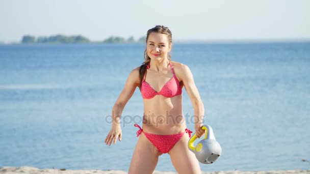 Beautiful, athletic, sexy young woman in swimsuit, coach, instructor, performs, doing exercises with weights. On the beach, near the sea, river, in summer, in sun rays. — Stock Video