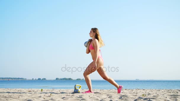 Beautiful, athletic, sexy young woman in swimsuit, coach, instructor, performs, doing exercises with weights. On the beach, near the sea, river, in summer, in sun rays. — Stock Video