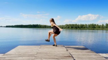 Zıplama, germe, farklı egzersizleri yapıyor güzel, atletik genç sarışın bir kadın lunges, ağız kavgası. Göl, nehir, mavi gökyüzü ve orman arka planda, yaz güneşli gün.
