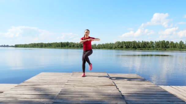 Vacker, atletisk ung blond kvinna stretching, göra olika övningar, hoppning, utfall, knäböj. Sjön, floden, blå himmel och skogen i bakgrunden, solig sommardag. — Stockvideo