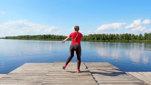 Mooie, sportieve jonge blonde vrouw uitrekken, doen verschillende oefeningen, springen, lunges, squats. Meer, rivier, blauwe lucht en bos op de achtergrond, zonnige zomerdag. — Stockvideo