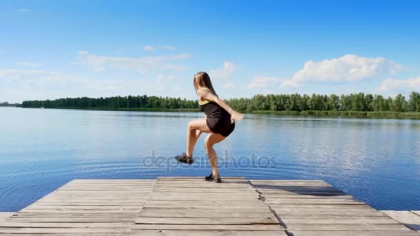 Hermosa, atlética joven rubia estirándose, haciendo diferentes ejercicios, saltos, embestidas, sentadillas. Lago, río, cielo azul y bosque en el fondo, día soleado de verano . — Vídeos de Stock