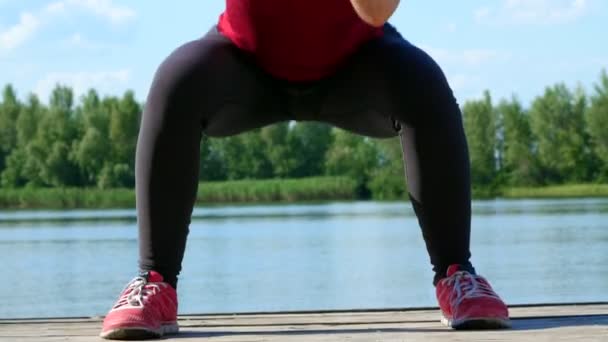 Hermosa, atlética joven rubia estirándose, haciendo diferentes ejercicios, saltos, embestidas, sentadillas. Lago, río, cielo azul y bosque en el fondo, día soleado de verano . — Vídeo de stock