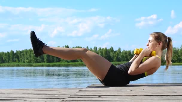 Mooie, sportieve jonge blonde vrouw uitrekken, swings pers. Meer, rivier, blauwe lucht en bos op de achtergrond, zonnige zomerdag — Stockvideo