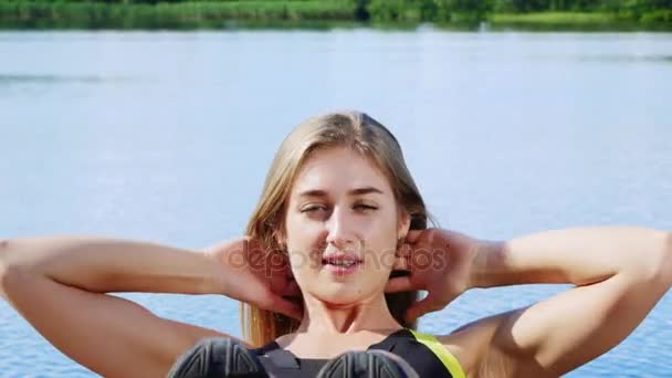 Hermosa, atlética joven rubia estirándose, columpios prensa. Lago, río, cielo azul y bosque en el fondo, día soleado de verano. primer plano — Vídeos de Stock