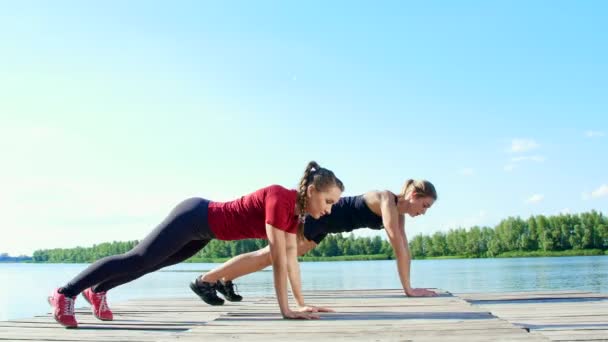 Dos hermosas mujeres rubias atléticas, haciendo ejercicios de coordinación y equilibrio sincrónicamente. Lago, río, cielo azul y bosque en el fondo, día soleado de verano — Vídeos de Stock