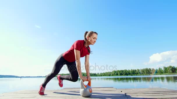 Bella, atletica giovane donna bionda che fa diversi esercizi con pesi, manubri, affondi, squat. Lago, fiume, cielo blu e foresta sullo sfondo, giornata estiva soleggiata — Video Stock