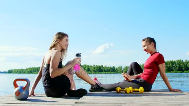 To vakre, atletiske, blonde jenter, kvinner hviler etter trening utendørs, drikker vann, snakker, ler. Innsjø, elv, blå himmel og skog i bakgrunnen, sommersoldag – stockvideo