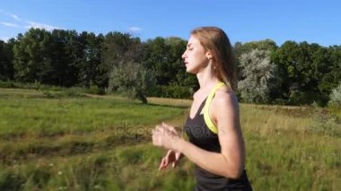 Güzel, yüksek, atletik sarışın genç kadın, kız, üst ve şort bir koşu yapar, günbatımı, güneş ışınları yakınındaki orman, mavi gökyüzü, yeşil çim arasında çayır yol boyunca çalışır