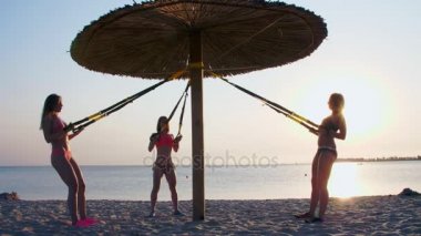 fitness trx sistemi, Trx askı kayışları ile egzersizleri yapıyor üç atletik, seksi genç kadınlar mayo, eğitmenler. Kumsalda, yaz, günbatımı, güneş ışınları.