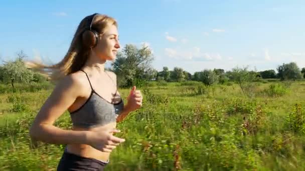 Vacker, hög, atletisk blond ung kvinna, tjej sätter hörlurarna och börjar köra, i topp och shorts gör en joggingtur, längs äng Stig, nära skogen, i solnedgången, i solens strålar — Stockvideo