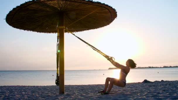 Athletische, sexy junge Frau im Badeanzug, Trainer, Instruktor, führt Übungen mit Fitness-Trx-System, Trx-Aufhängebändern durch. am Strand, Sommer, Sonnenuntergang, in der Sonne. — Stockvideo