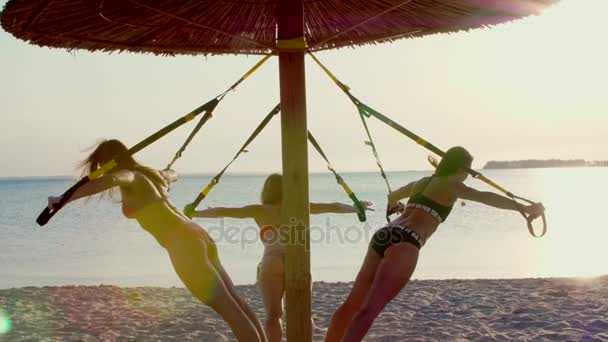 Drie atletisch, sexy jonge vrouwen in zwemkleding, instructeurs, doen oefeningen met fitness trx systeem, Trx suspension bandjes. Op het strand, in de zomer, bij zonsondergang, in zonnestralen. — Stockvideo